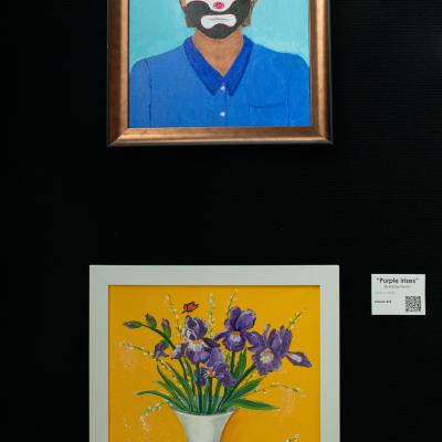 A painting of a clown and a painting of flowers in a vase hang on a black wall.