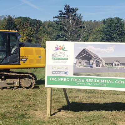 An architectural drawing depicts the future Frese center, with a yellow excavating machine parked behind the sign.